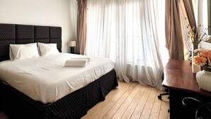 A bedroom with a large bed, wooden flooring, a desk, and a window with curtains. - Peaceful 2 minutes European Parliament (Brussels)