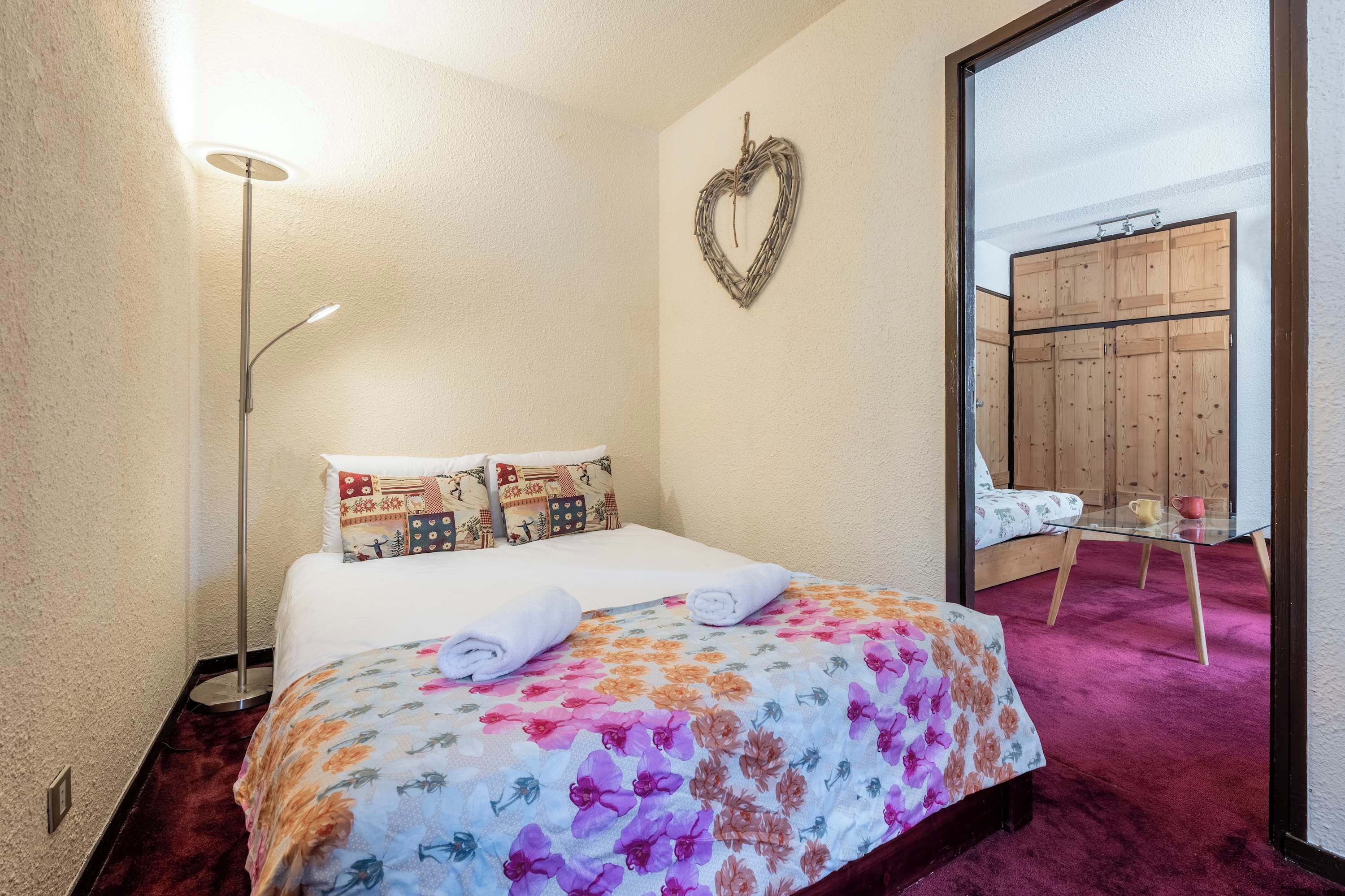 A hotel room with a bed, a heart-shaped decoration on the wall, a wooden wardrobe, and a small table with a chair.