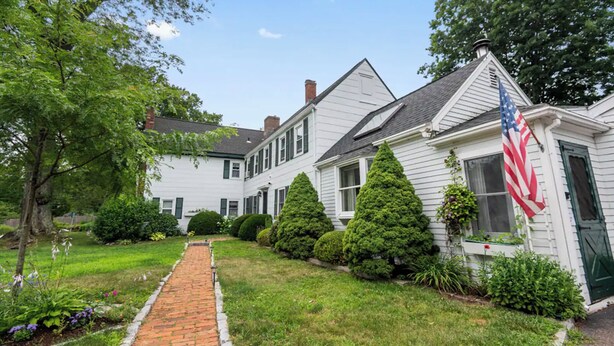 A two-story white house with a brick pathway leading to the front door, surrounded by lush greenery and a small American flag. - Great Space to be Together and Spread out (Hamilton)