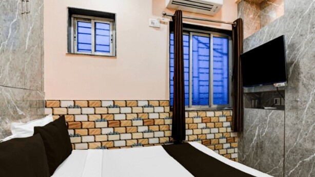 Une chambre d’hôtel avec un mur décoré d’un motif en briques, un lit avec une literie blanche et noire, une petite fenêtre avec des rideaux et un téléviseur à écran plat fixé au mur.