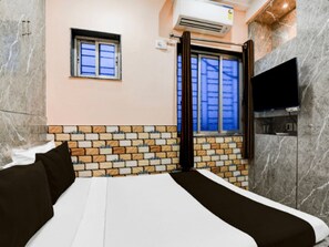 Une chambre d’hôtel avec un mur décoré d’un motif en briques, un lit avec une literie blanche et noire, une petite fenêtre avec des rideaux et un téléviseur à écran plat fixé au mur.