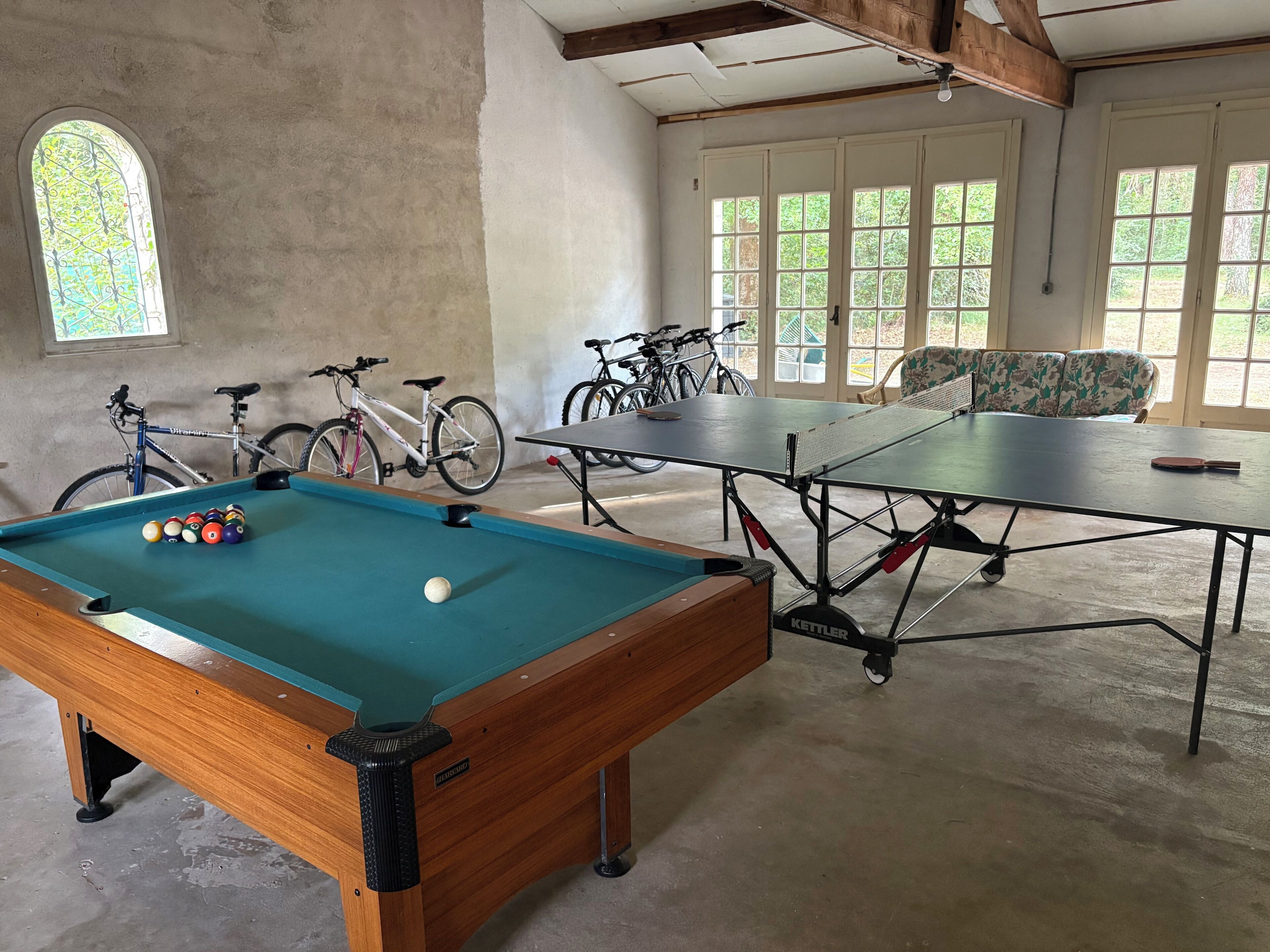 A pool table with balls, a billiards table, bicycles, and a sofa in a room with large windows and a concrete floor.