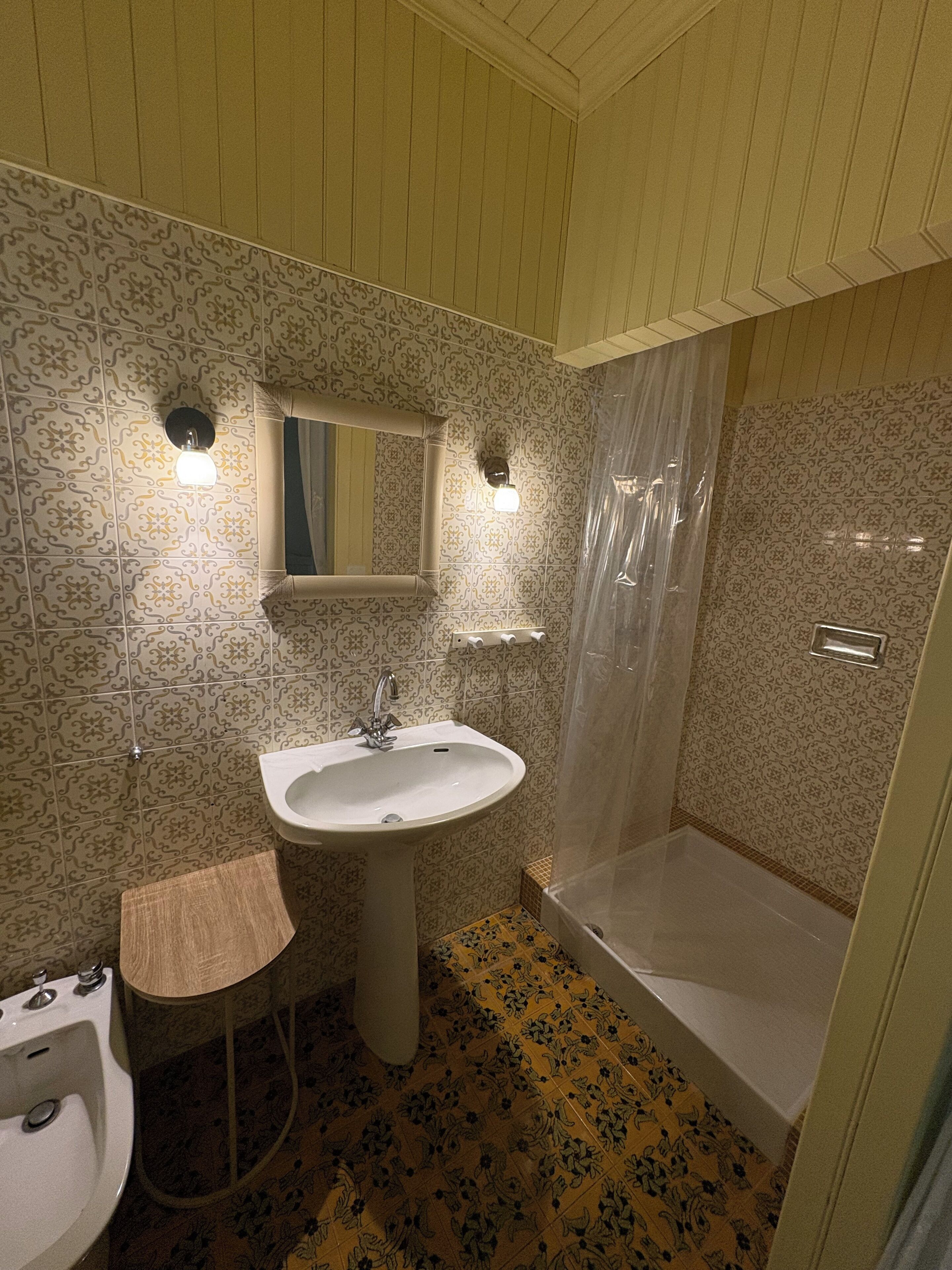 Un baño con baldosas estampadas, un lavamanos pedestal, un espejo, un cerramiento de ducha y un taburete pequeño.