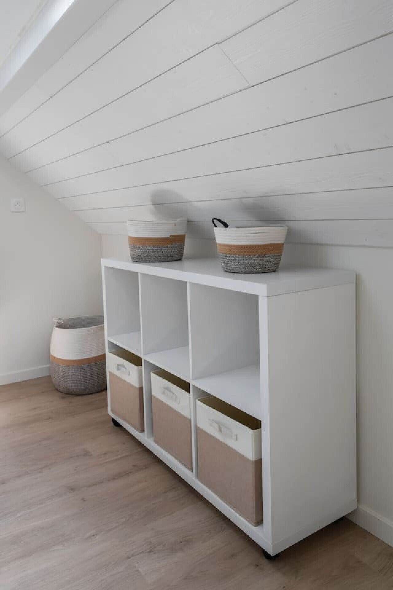 A white storage unit with drawers and shelves, placed against a white wall in a room with wooden flooring.