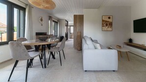 En moderne stue med sofa, spisebord og stole. Der er et stort vindue, et fjernsyn og en dekorativ vase.