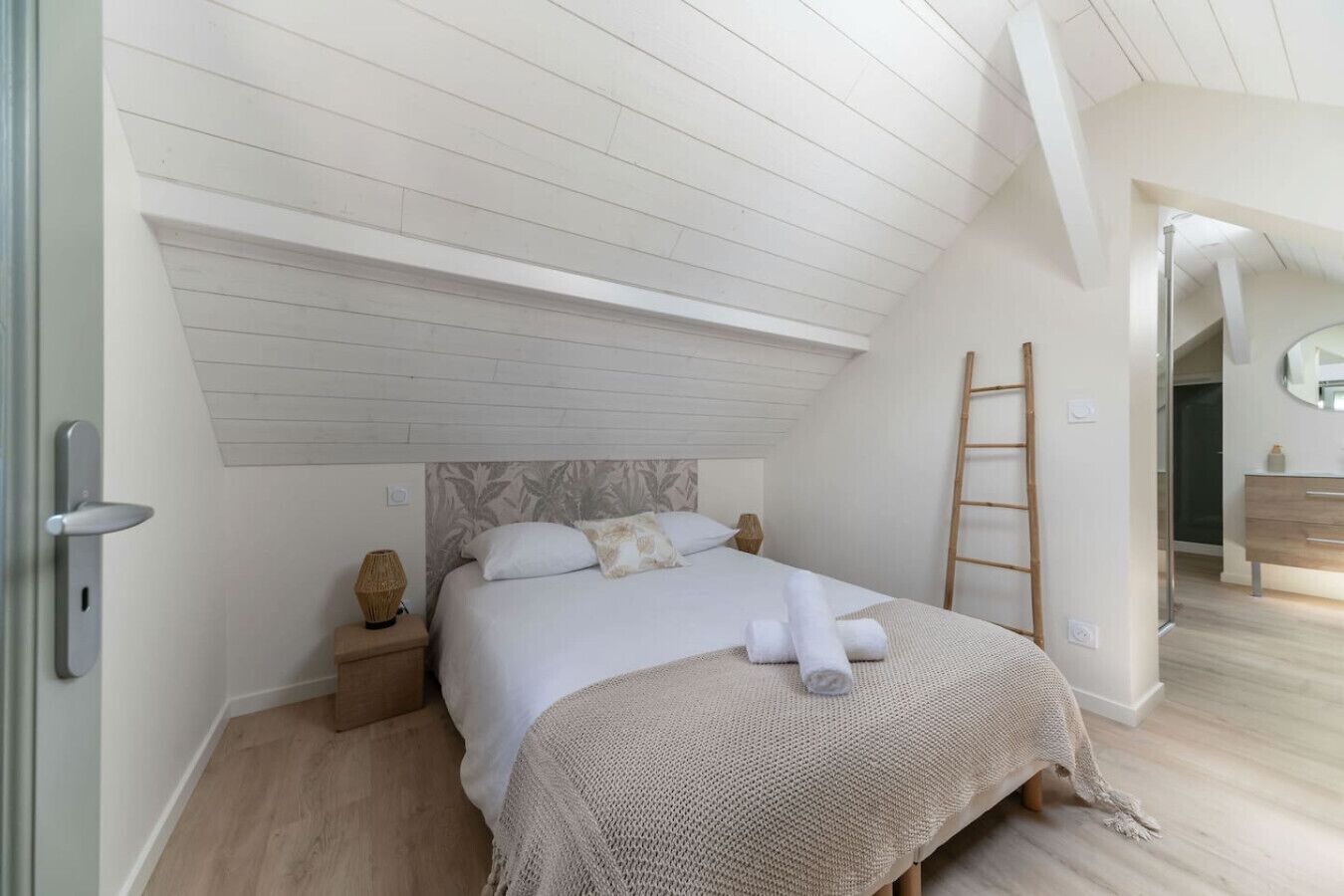 Une chambre avec un plafond en pente, un lit recouvert d’une courtepointe blanche, une échelle en bois et une table de chevet avec une lampe.
