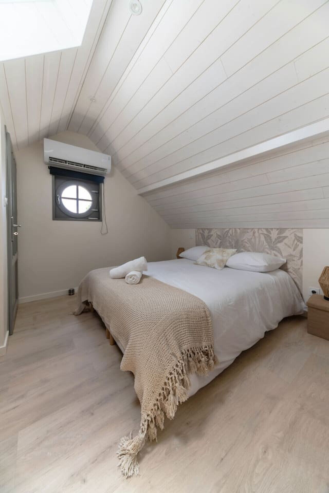Een slaapkamer met een schuine plafond, een bed met een beige deken, een klein raam en een airconditioning.