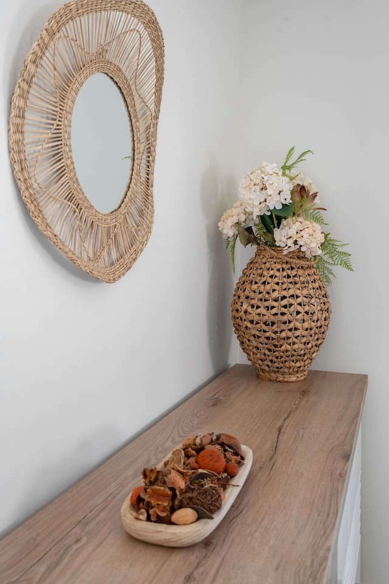 Una pared con un espejo tejido y un jarrón decorativo con flores sobre una repisa de madera.