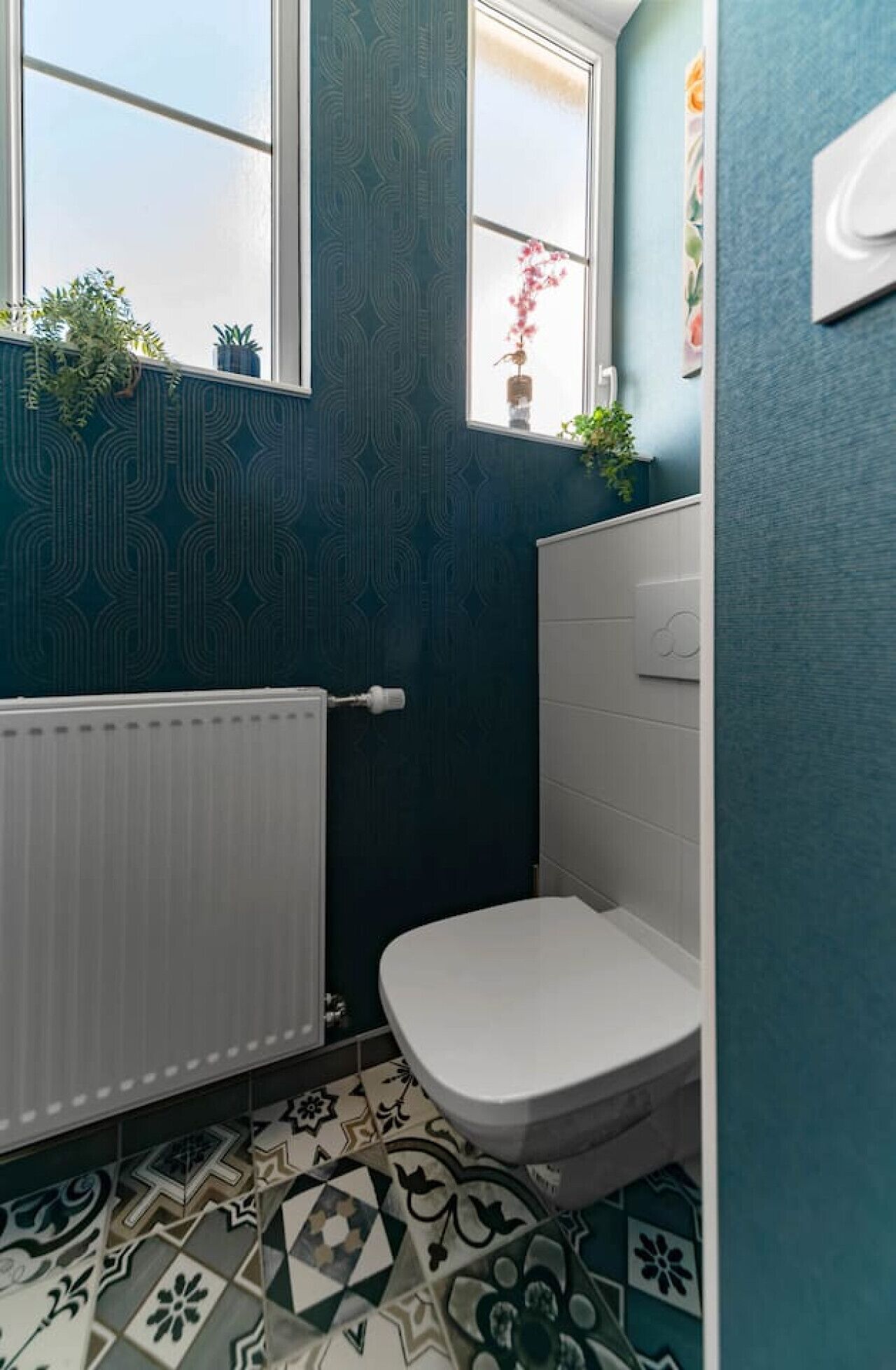 Un baño con inodoro blanco, baldosas de piso estampadas, un radiador y una ventana con plantas en el alféizar.