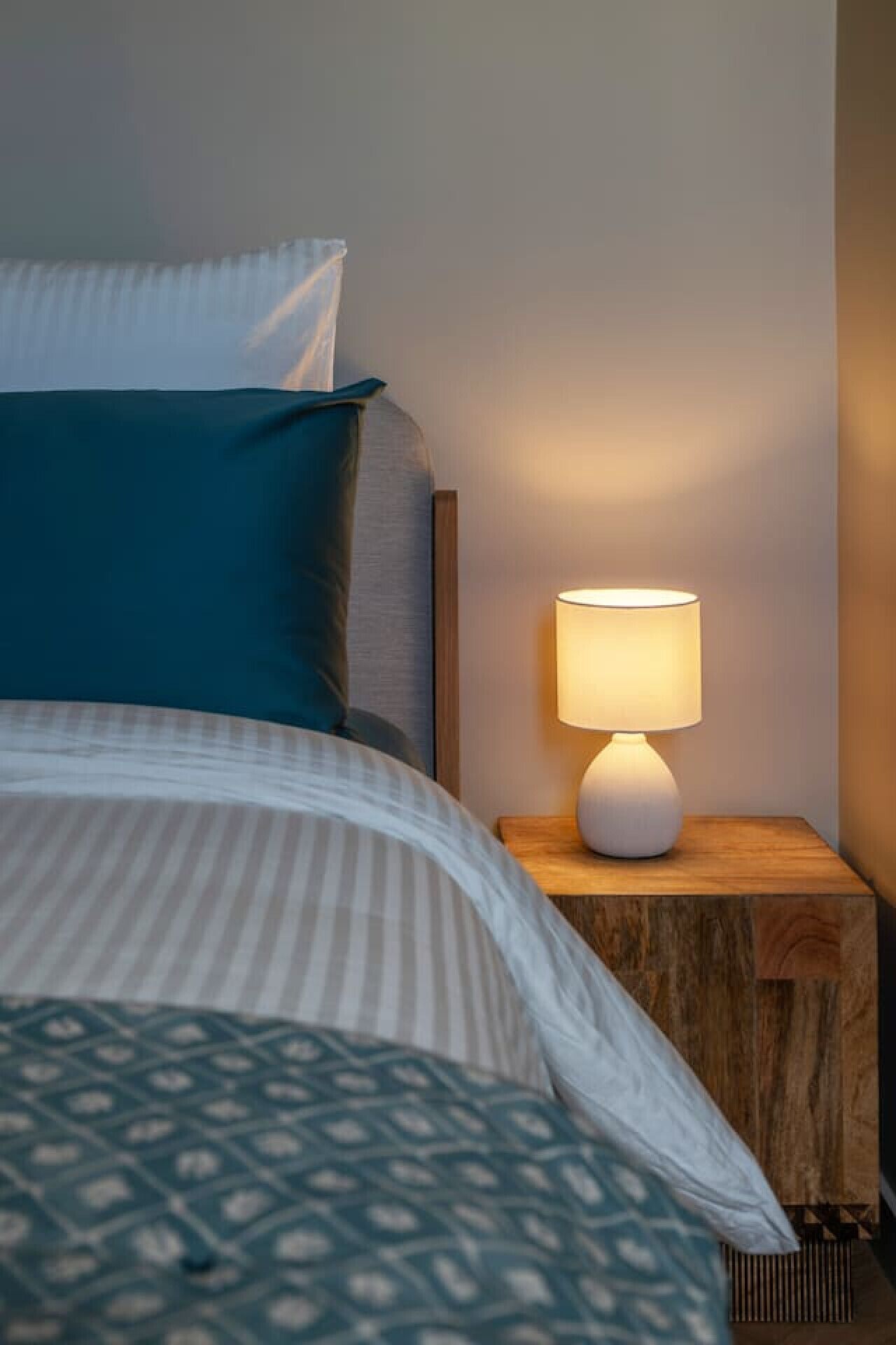 Een net opgemaakt bed met gestreepte beddengoed en een decoratieve plaid, een bedlamp op een houten nachtkastje, en een muur met een structuur.