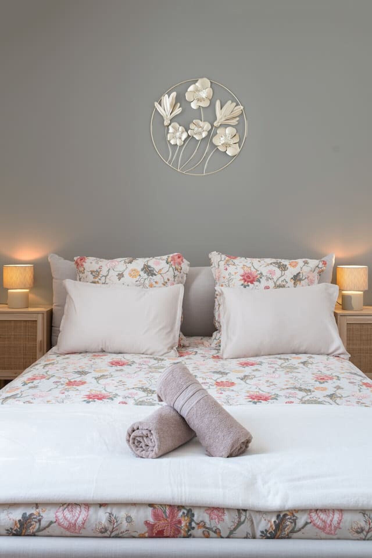 A neatly made bed with floral bedding, two matching pillows, a rolled towel, and a wall-mounted floral decoration.