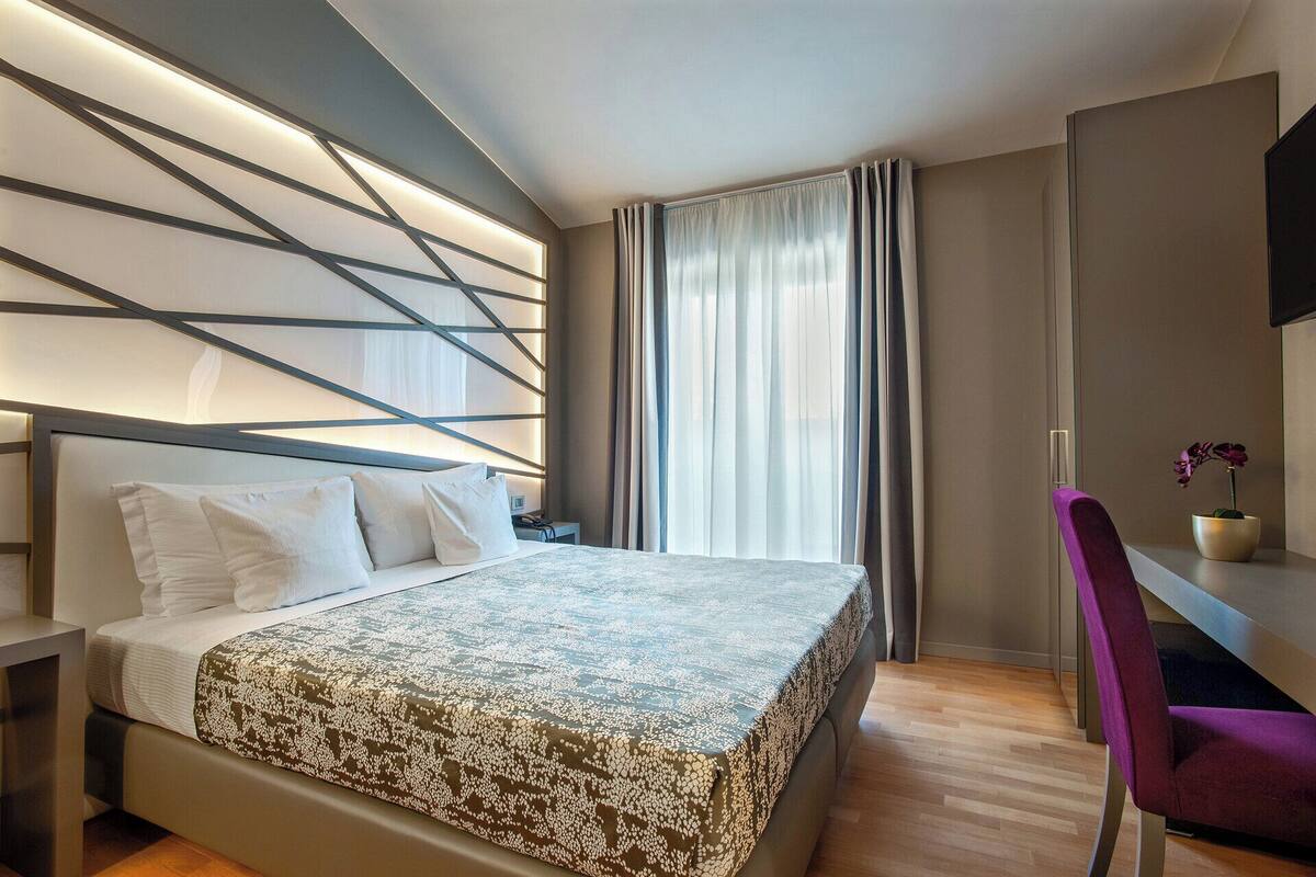 A modern hotel room with a large bed, a desk with a chair, and a window with curtains.