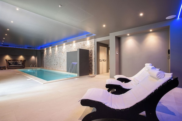 Un área de piscina interior moderna con una pared de piedra, una silla de descanso y una puerta que da a un vestuario.