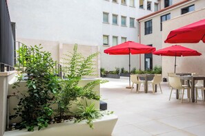 Un patio al aire libre con mesas y sillas, sombrillas rojas y plantas en macetas.