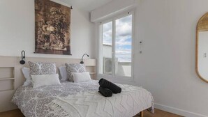Ein Schlafzimmer mit einem Bett, einem großen Fenster, einer Hängelampe und einem an der Wand befestigten Spiegel.