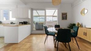 Eine moderne Küche mit einer zentralen Kochinsel, einem Essbereich mit rundem Tisch und Stühlen sowie einem Ausblick durch ein großes Fenster.