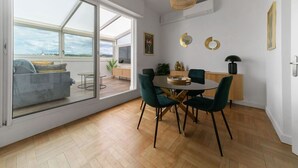 Ein modernes Wohnzimmer mit einer Glastür zum Balkon, einem runden Esstisch mit Stühlen und einer Küchenzeile mit Holzschrank und Pendelleuchte.