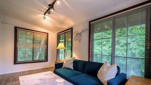 A living room with a dark green sofa, two side tables, a lamp, and large windows with blinds.