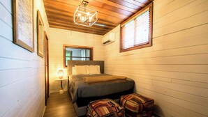 A narrow, cozy room with a wooden ceiling, a bed with a gray blanket, two patterned ottomans, a small table with a lamp, and a window with blinds.