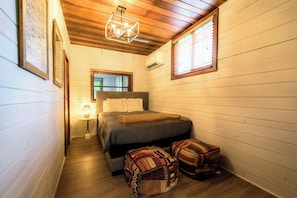 Una habitación estrecha con techo de madera, una cama con colcha gris, dos otomanas estampadas, una mesita de noche con lámpara y una ventana con persianas.
