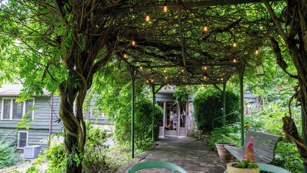 Un espace extérieur aménagé, abrité et doté de tables et de chaises en bois, entouré d’une végétation luxuriante et agrémenté d’une pergola illuminée par des guirlandes lumineuses.