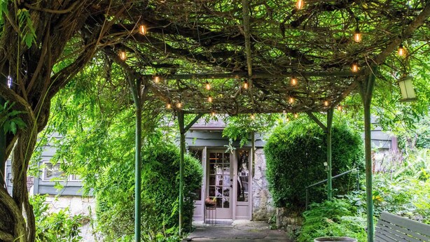 A covered garden path leading to a building entrance, surrounded by lush greenery and a wooden bench. - Stone & Timber Studio Into the Woods Near Downtown (Hendersonville)