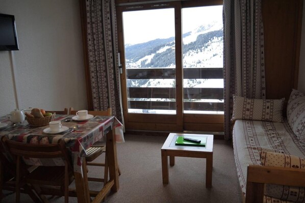 Une pièce chaleureuse avec une table à manger dressée pour deux personnes, un canapé et une vue sur des montagnes enneigées, visible à travers une grande fenêtre.