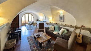 A spacious loft with a high ceiling, a cozy living area with a sofa and coffee table, a dining area with chairs, and a kitchen in the background.