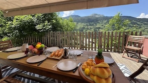 An outdoor dining table set with plates, cutlery, bread, and fruit, overlooking a mountainous landscape.