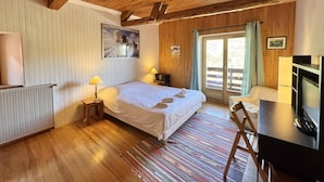 A cozy bedroom with a wooden ceiling, a bed with a white duvet, a small bedside table with a lamp, a television, and a window with curtains.
