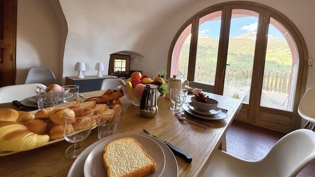 A dining table set with bread, fruits, and glasses in a room with a large arched window.
