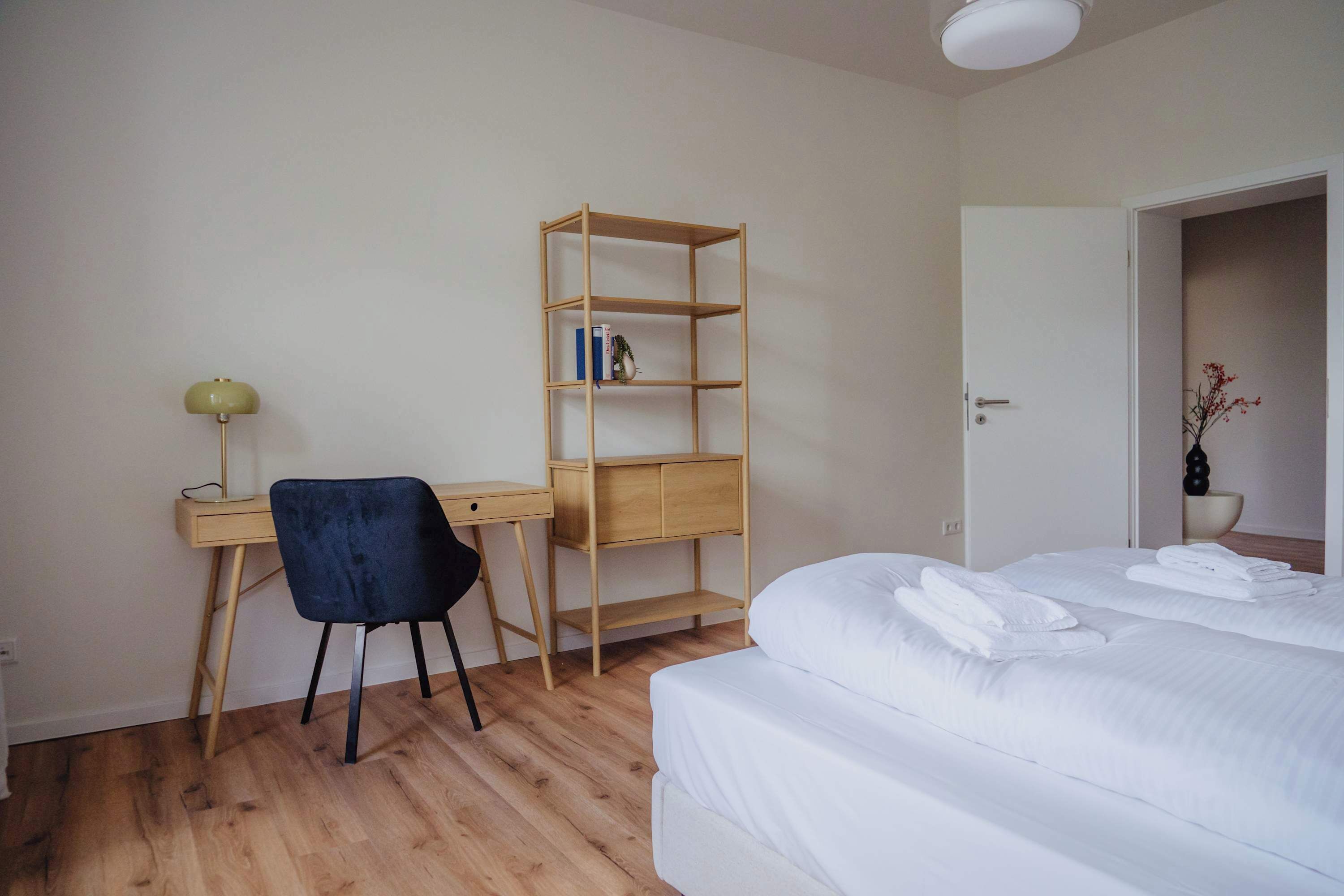 Une chambre à coucher comprenant un lit, un bureau avec une chaise, une bibliothèque en bois et une porte donnant sur une autre pièce.