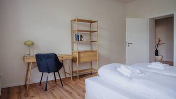A bedroom with a bed, a desk with a chair, a wooden bookshelf, and a door leading to another room.