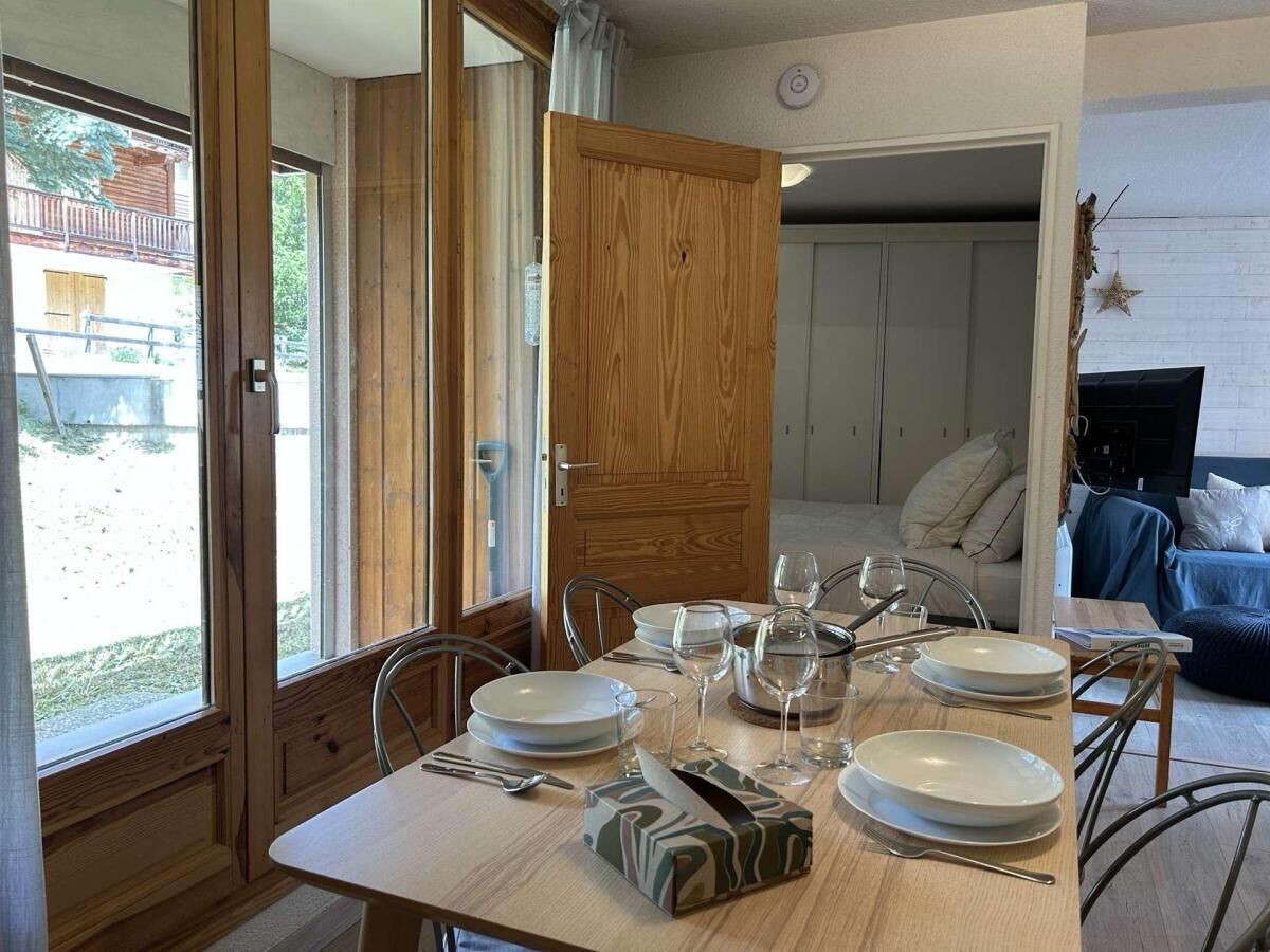 A dining area with a wooden table set for four, surrounded by chairs. A wooden door is open, revealing a living area with a sofa and a TV.