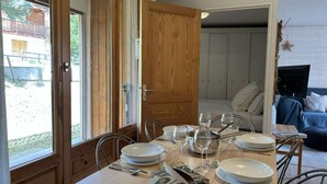 A dining area with a wooden table set for four, surrounded by chairs. A wooden door is open, revealing a living area with a sofa and a TV.