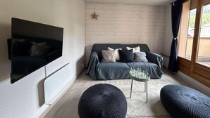 A modern living room with a flat-screen TV mounted on the wall, a dark-colored sofa with cushions, a glass coffee table, and two round ottomans.