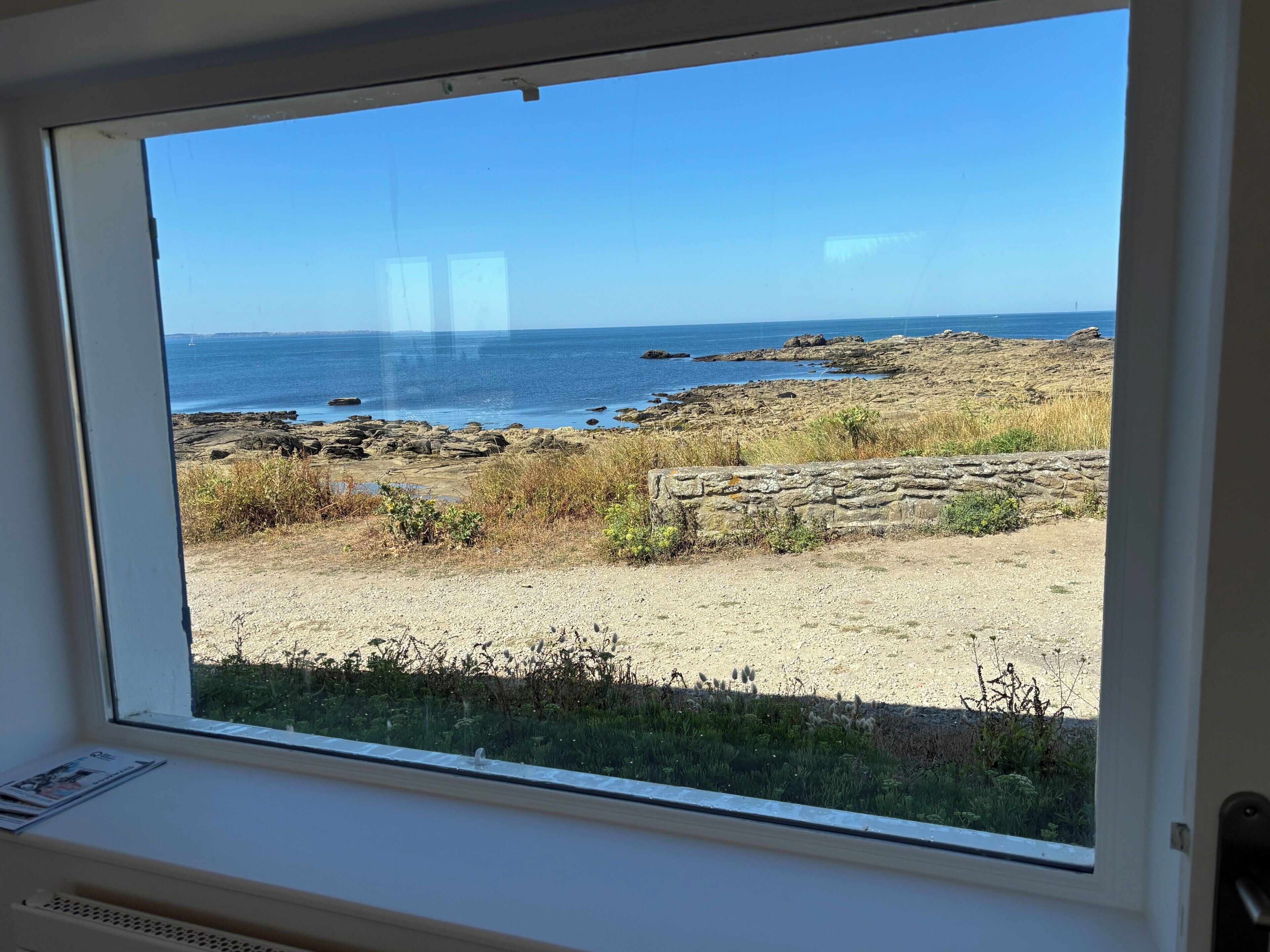 A window view of a rocky beach with the sea and a clear sky.
