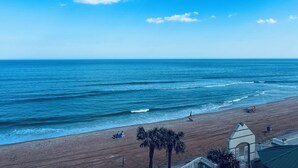 A beach with a clear sky, palm trees, and a building with a pointed roof. - ~The Shoreline~ Oceanfront Studio Condo (Daytona Beach)