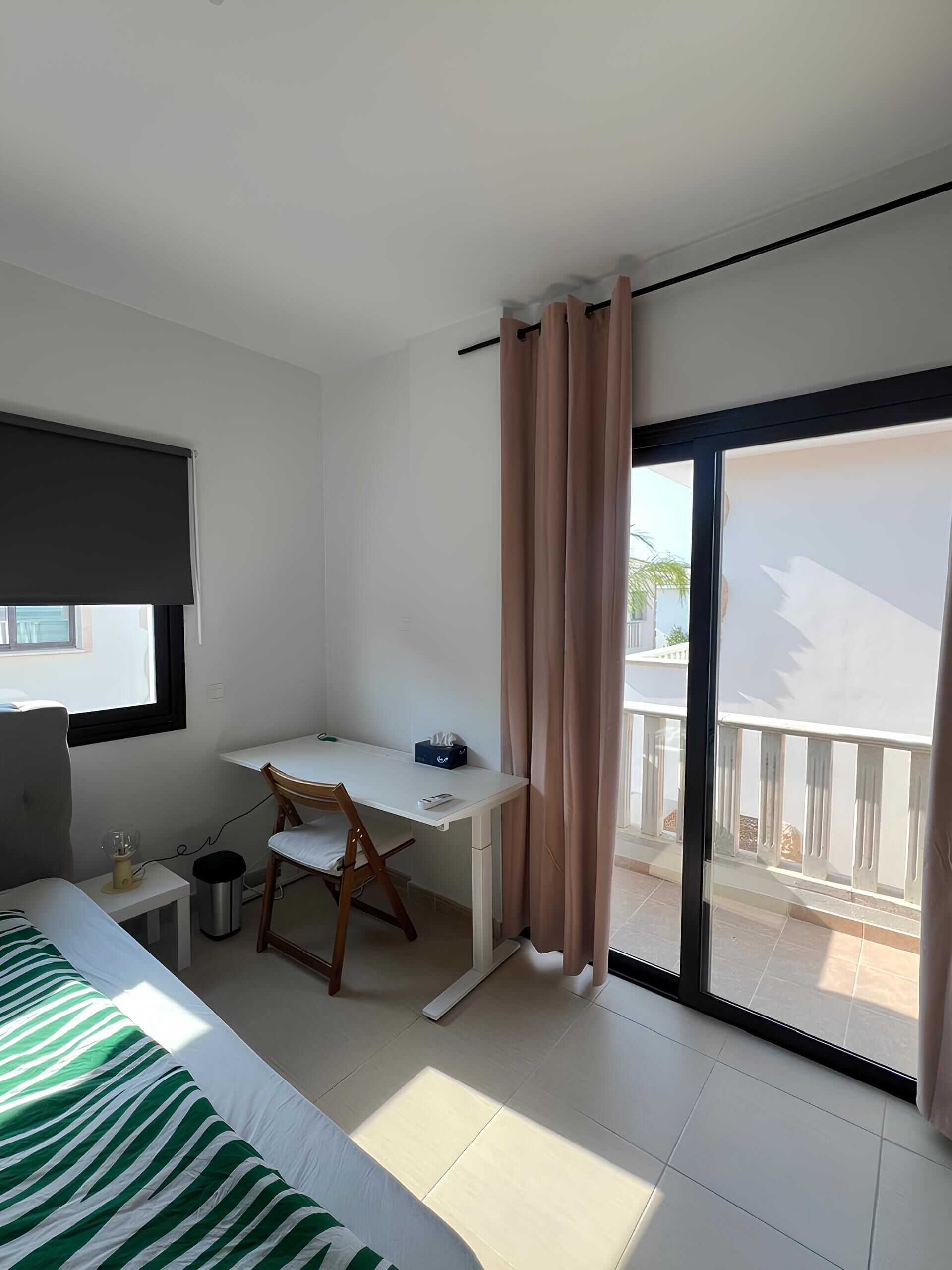 Habitación de hotel con una cama, un escritorio, una silla, una mesita, un cesto de basura y una ventana con persiana.