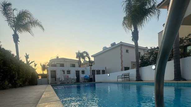 Una piscina con agua cristalina, rodeada por un sendero pavimentado y palmeras.