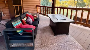 Eine Holzhütte mit überdachter Veranda, einer schwarzen Couch mit bunten Kissen, einem kleinen Tisch und Blick auf Herbstbäume und Berge.