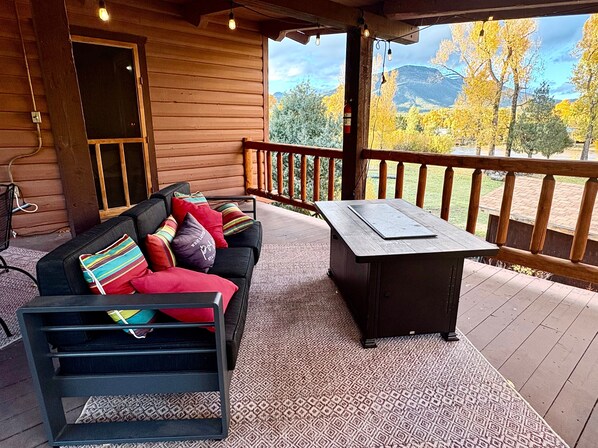 A wooden cabin with a covered porch, a black sofa with colorful cushions, a small table, and a view of autumn trees and mountains.