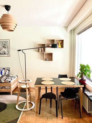 A dining area with a wooden table, black chairs, and a floor lamp. There are shelves with decorative items and a plant by the window.