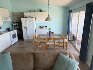 Une cuisine compacte équipée d’un four à micro-ondes, d’une cuisinière et d’un réfrigérateur, un coin repas avec une table et des chaises en verre, et un canapé avec des coussins.
