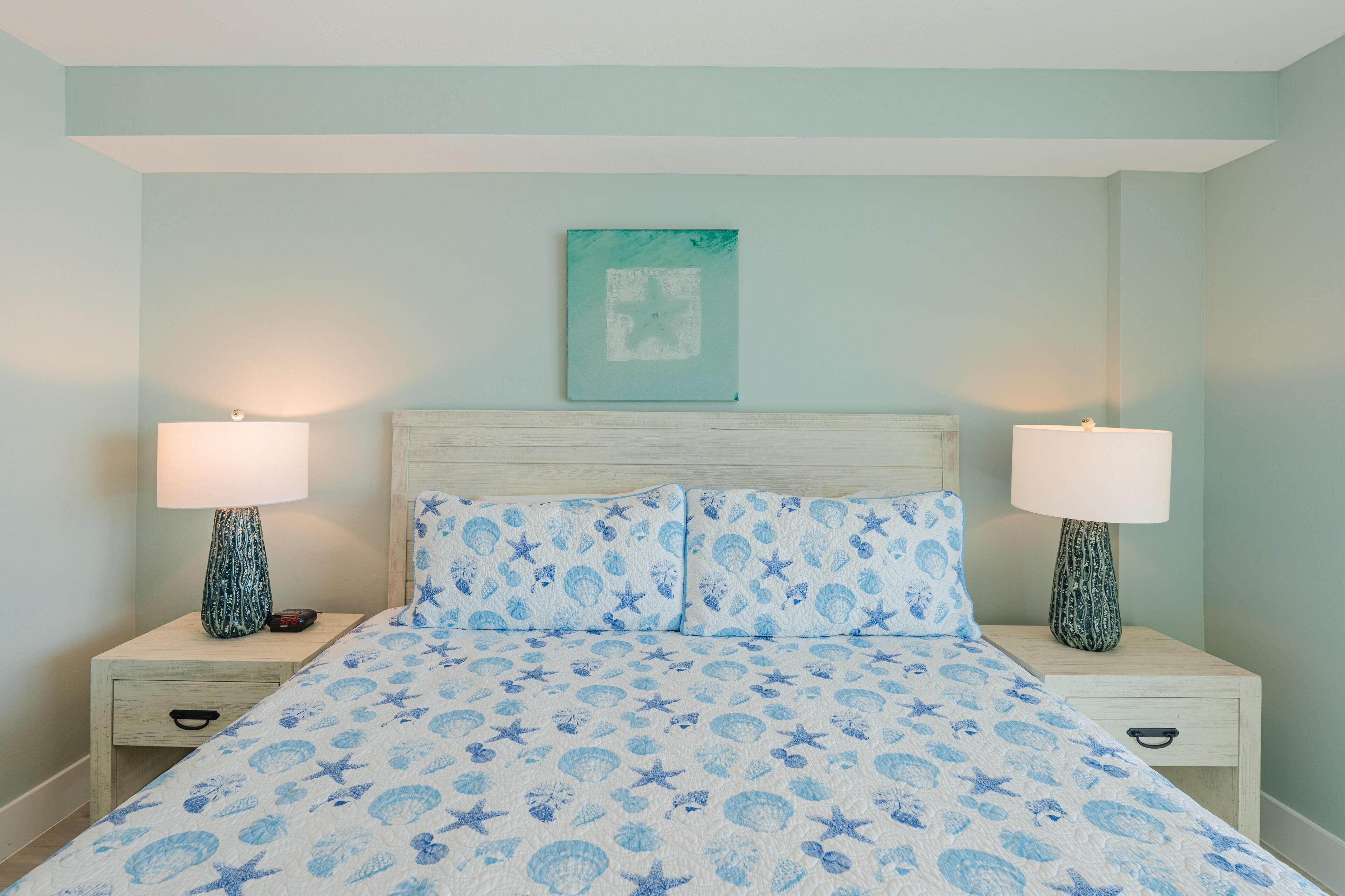 Una cama con una colcha azul y blanca estampada, dos mesitas de noche con lámparas, y una obra de arte enmarcada en la pared.