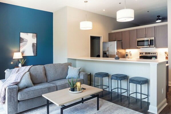 Una sala de estar moderna con un sofá gris, una mesa de centro y una zona de cocina con un taburete de bar y un microondas.
