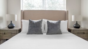A neatly made bed with two gray pillows, a white pillow, and a beige headboard. There are two bedside tables with lamps and a window with blinds. - North Concord Apartments By Landing (Concord)