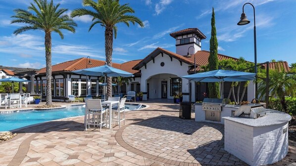 A pool area with lounge chairs and umbrellas, a barbecue station, and a building with a tiled roof and a clock tower.