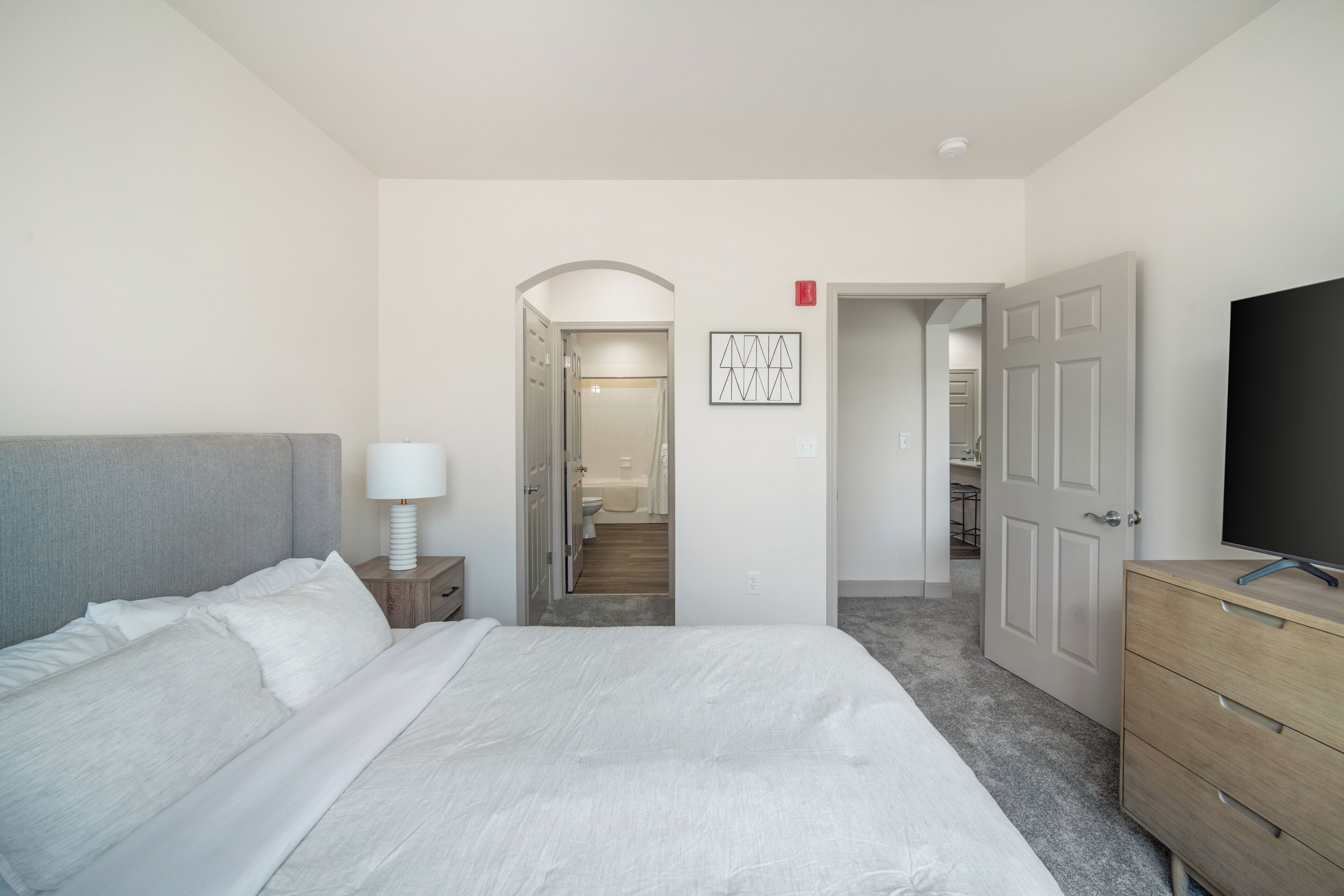 A bedroom with a bed, a nightstand, a dresser, and a TV. There is a door leading to a bathroom and a hallway.