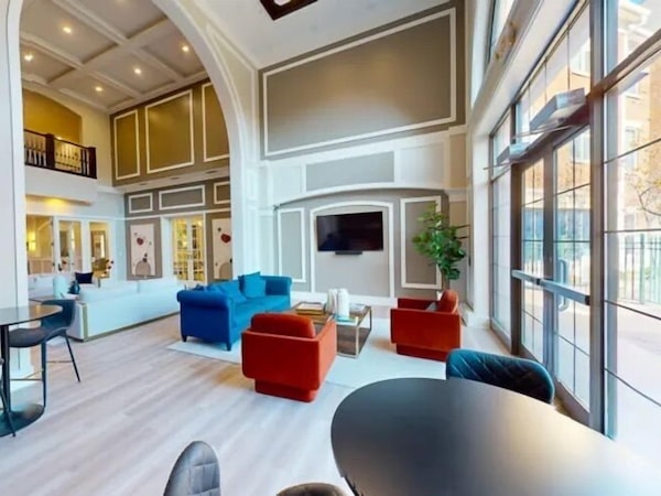 A spacious living area with a high ceiling, large windows, and a fireplace.